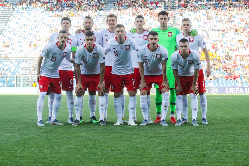 Reprezentacja Polski do lat 21 przed meczem z rówieśnikami z Belgii - pierwszym spotkaniem na Euro 2019. Polacy w białych koszulkach z czerwonym kołnierzem, w czerwonych spodenkach. Bramkarz Grabara w jaskrawozielonej bluzie.