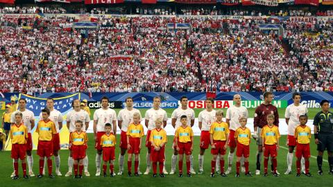 Reprezentacja Polski na środku boiska. Polacy w białych koszulkach, czerwonych spodenkach. Bramkarz Artur Boruc w bordowej bluzie. Przed piłkarzami stoją dzieci ubrane w żółte koszulki i czerwone spodenki.